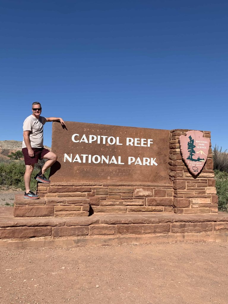 Me standing at the Capitol Reef National Park Entrance sign against a bright blue sky.